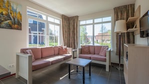 Una sala de estar moderna con dos sofás, una mesa de centro y un televisor sobre un mueble bajo. Amplias ventanas ofrecen luz natural y vistas al exterior.