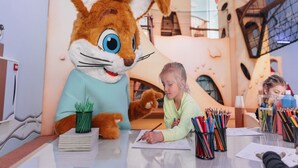 Un niño dibujando en una mesa con un gran conejo como mascota y diversos materiales de arte.