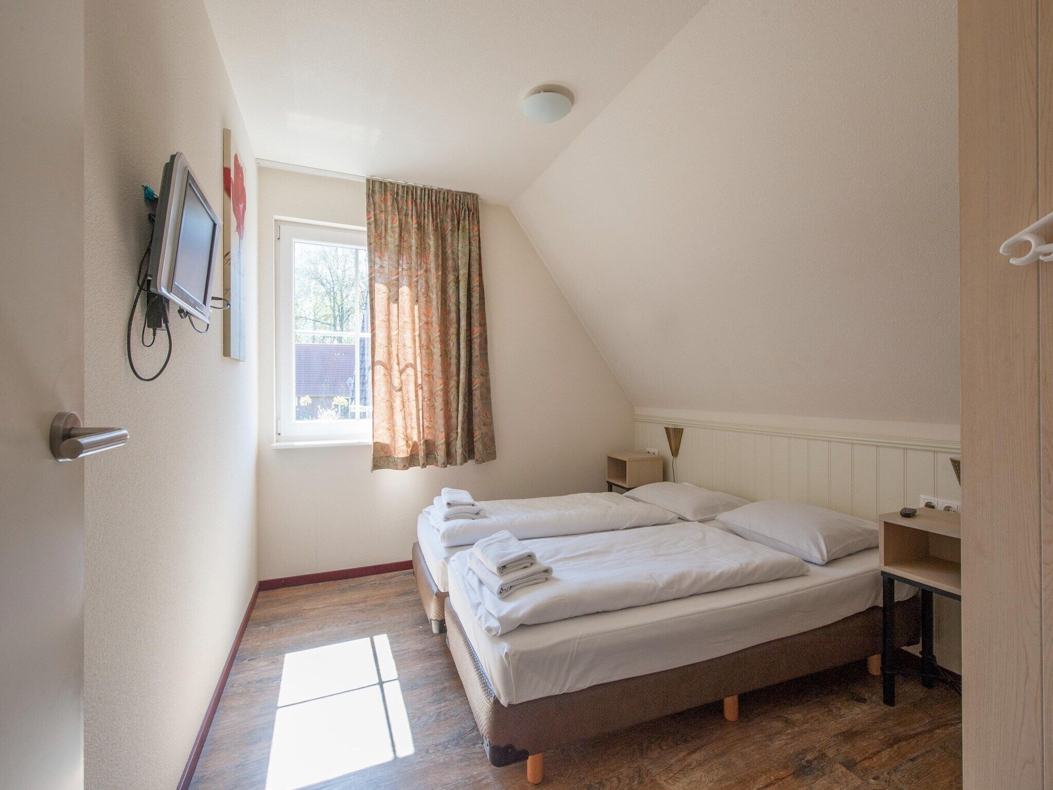 Ein Schlafzimmer mit Bett, Vorhängen am Fenster, einem Nachttisch und einem an der Wand montierten Fernseher.