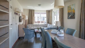 Una sala de estar moderna con sofá, mesa de comedor y sillas. Hay un televisor montado en la pared, una ventana con cortinas y un cuadro.