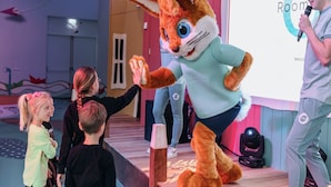 Una persona disfrazada de conejo en un escenario, con un micrófono, una pantalla de proyector que muestra la palabra 'Room' y una plataforma de madera.