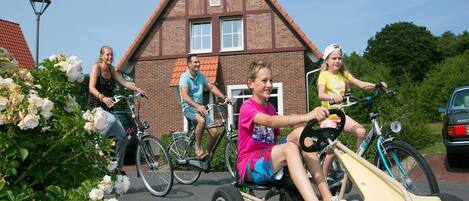 A group of people riding bicycles and a tricycle in a residential area with a brick house and blooming flowers.