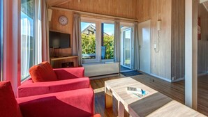 Ein modernes Zimmer mit Holzboden, einem Fernseher auf einer Holzablage, einem roten Sofa, einem Holztisch und großen Fenstern mit Vorhängen.