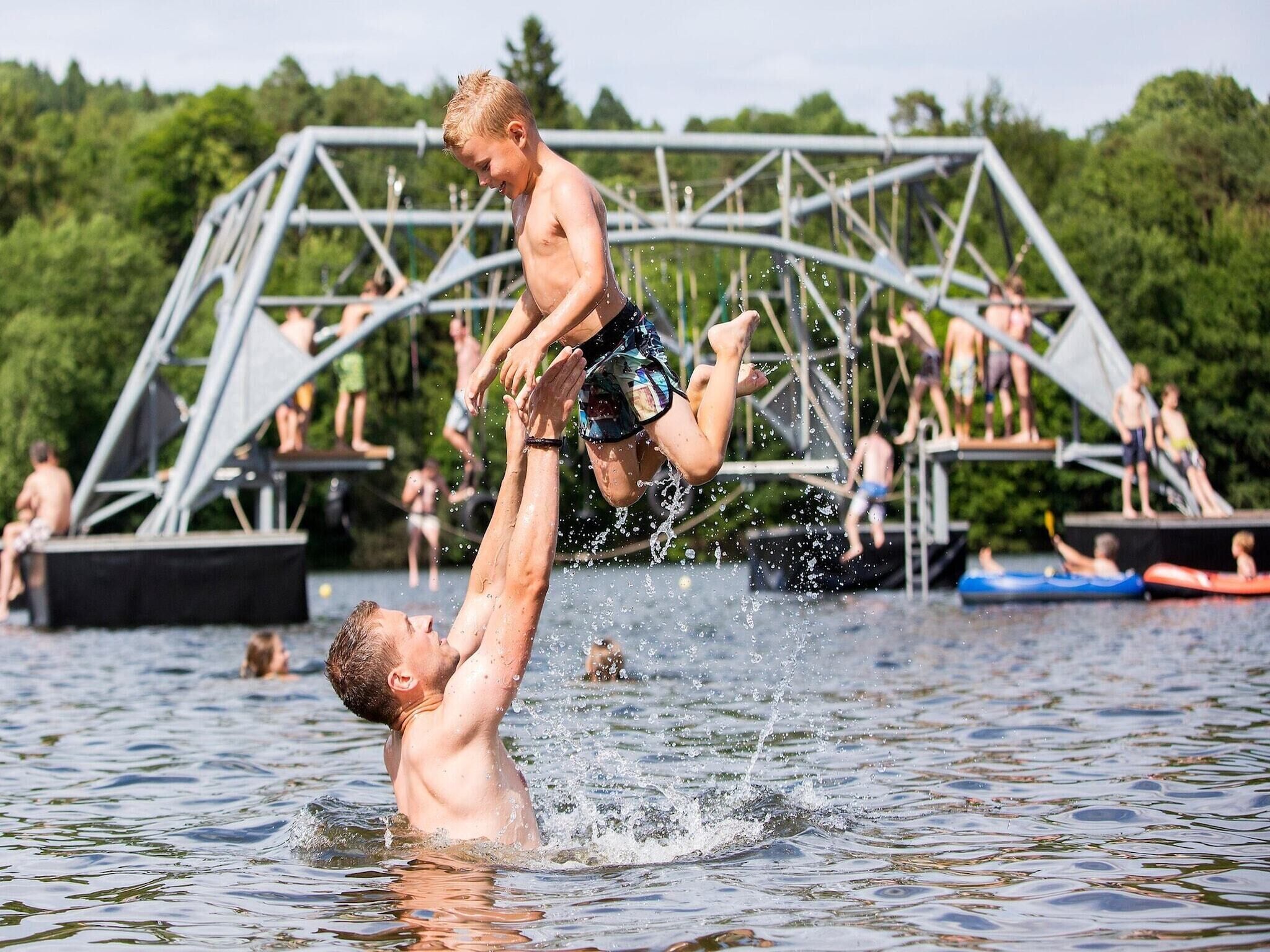 Un homme soulève un autre homme dans une étendue d’eau, avec une structure métallique en arrière-plan.