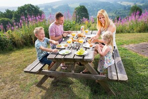 Une famille est assise à une table de pique-nique en bois, à l’extérieur, et profite d’un paysage magnifique en arrière-plan.