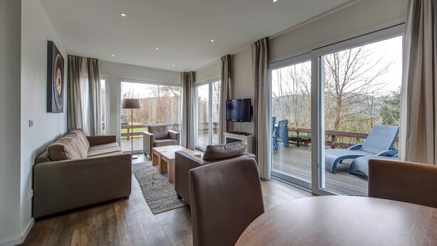 Un salon moderne comprenant un canapé, une table basse et un téléviseur, de grandes fenêtres et un balcon avec un espace salon extérieur.