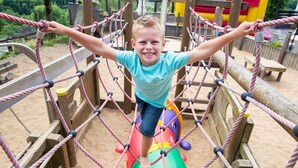Un enfant sur un pont de corde dans un espace de jeux extérieur, avec du sable et des structures en bois.