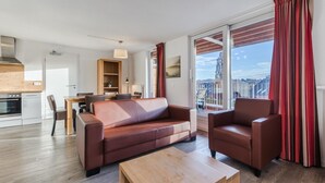 A modern living room with a sofa, armchair, and coffee table. There is a dining area with a table and chairs, and a kitchen with a stove. Large windows provide a view of the city. - Winterberg Stay with Beautiful View (Winterberg)