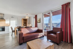 A modern living room with a sofa, armchair, and coffee table. There is a dining area with a table and chairs, and a kitchen with a stove. Large windows provide a view of the city. - Winterberg Stay with Beautiful View (Winterberg)