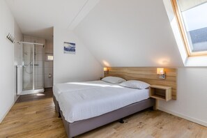 Ein Schlafzimmer mit Bett, Holz-Kopfstütze, Nachttisch, Wandleuchte, Dachfenster und Duschbereich.