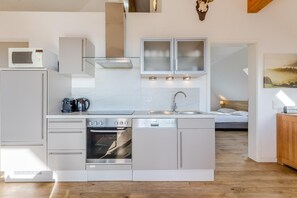 Una cocina moderna con horno empotrado, microondas y lavavajillas, suelo de madera y vistas a un dormitorio con cama y mesita de noche.