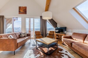 A modern loft apartment with a large window, a flat-screen TV, leather sofas, and a wooden coffee table.