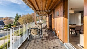 Una terraza de madera con mobiliario exterior, vista de los alrededores y entrada a un edificio.