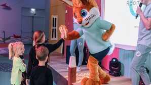 A person in a rabbit costume on a stage with a microphone, a projector screen displaying "Room", and a wooden platform. - Modern Bungalows by the Sea (De Koog)