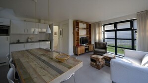 Een moderne keuken met witte kasten, een houten eettafel en een ingebouwde televisie. Er is een gezellige zithoek met een bank en een stoel, en een groot raam met gordijnen.