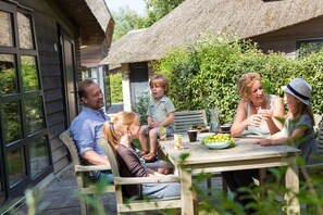 Una famiglia che cena all'aperto a un tavolo di legno, con un edificio a tetto di paglia sullo sfondo.