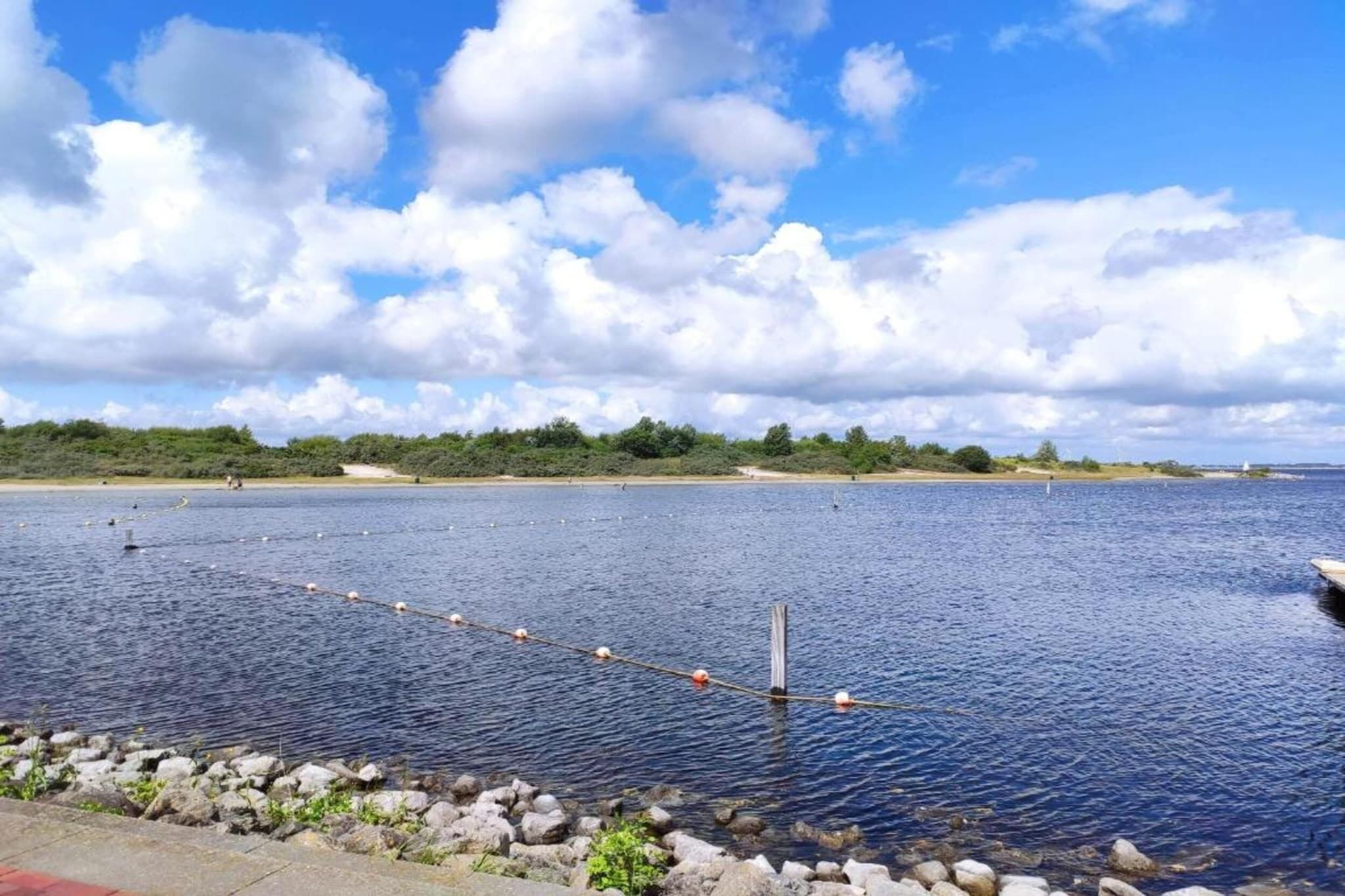 Ein ruhiger See mit Seilsperrung, felsigem Ufer und einer entfernten Baumgrenze unter teilweise bewölktem Himmel.