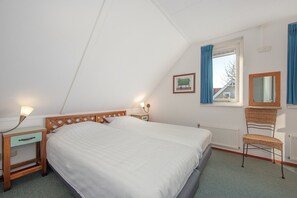 A bedroom with a large bed, a nightstand with a lamp, a chair, a window with curtains, and a framed picture on the wall.