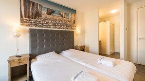 A hotel room with a large bed, a headboard, a nightstand, a bedside lamp, and a painting of a beach scene. - Modern Domburg Apartment (Domburg)