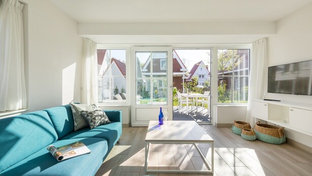 Ein helles Wohnzimmer mit einem türkisfarbenen Sofa, einem Couchtisch und einem Fernseher. Große Fenster bieten Blick auf Häuser und Bäume.