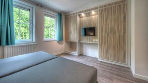A bedroom with a bed, a TV mounted on a wooden panel wall, and two windows with curtains.