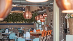 A modern restaurant with round tables and chairs, copper pendant lights, and a green wall with plants.