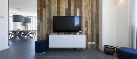 A modern living room with a large flat-screen TV mounted on a wooden wall, a white entertainment unit below, and a dining area in the background.