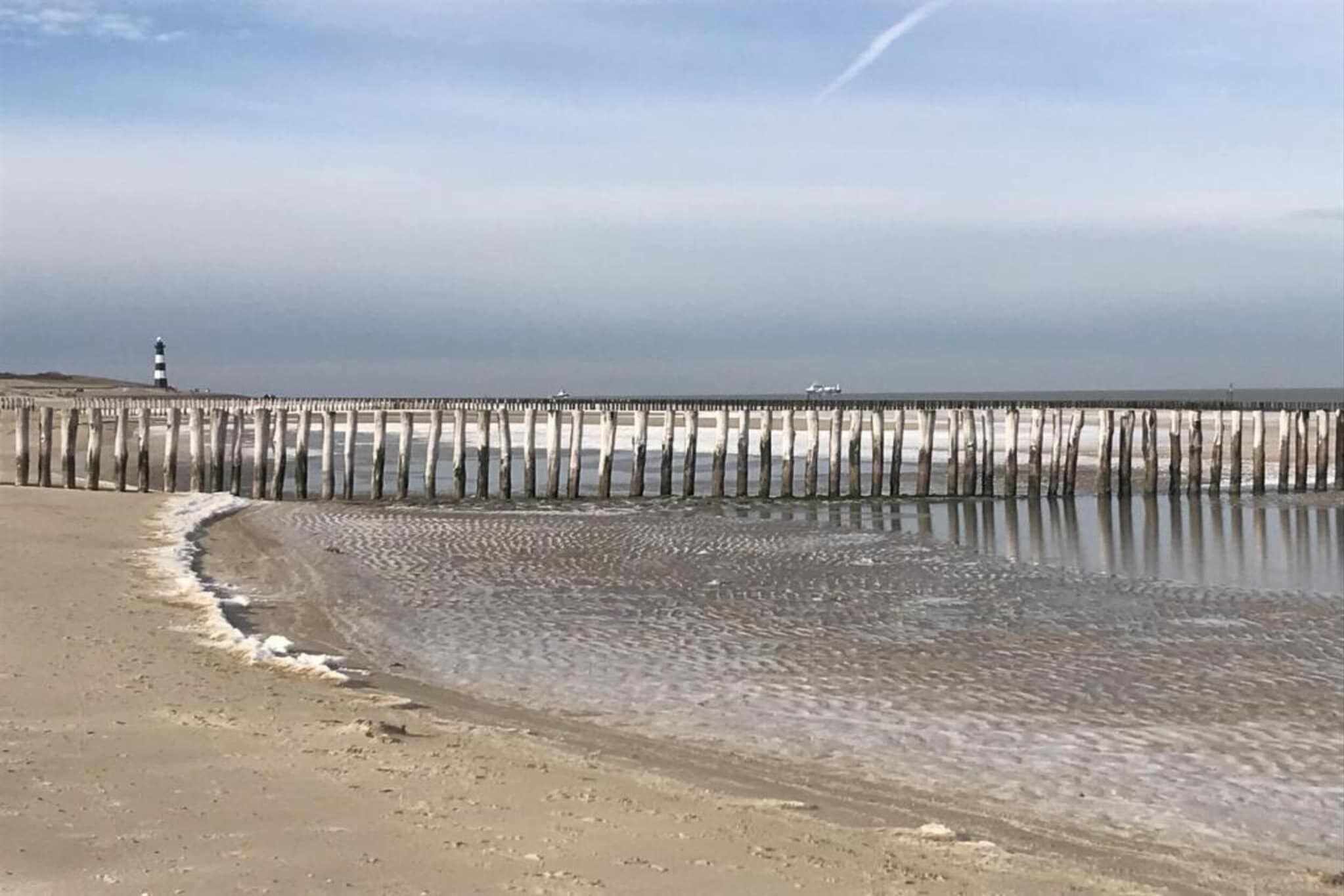 A beach with a wooden pier extending into the water, a lighthouse in the distance, and a clear sky.