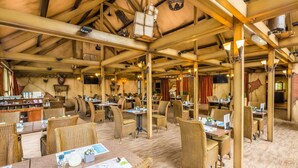 Ein Restaurant mit hohen Holzbalkendecken, Korbmöbeln und rustikalem Dekor, darunter Tierköpfe und eine Krone.