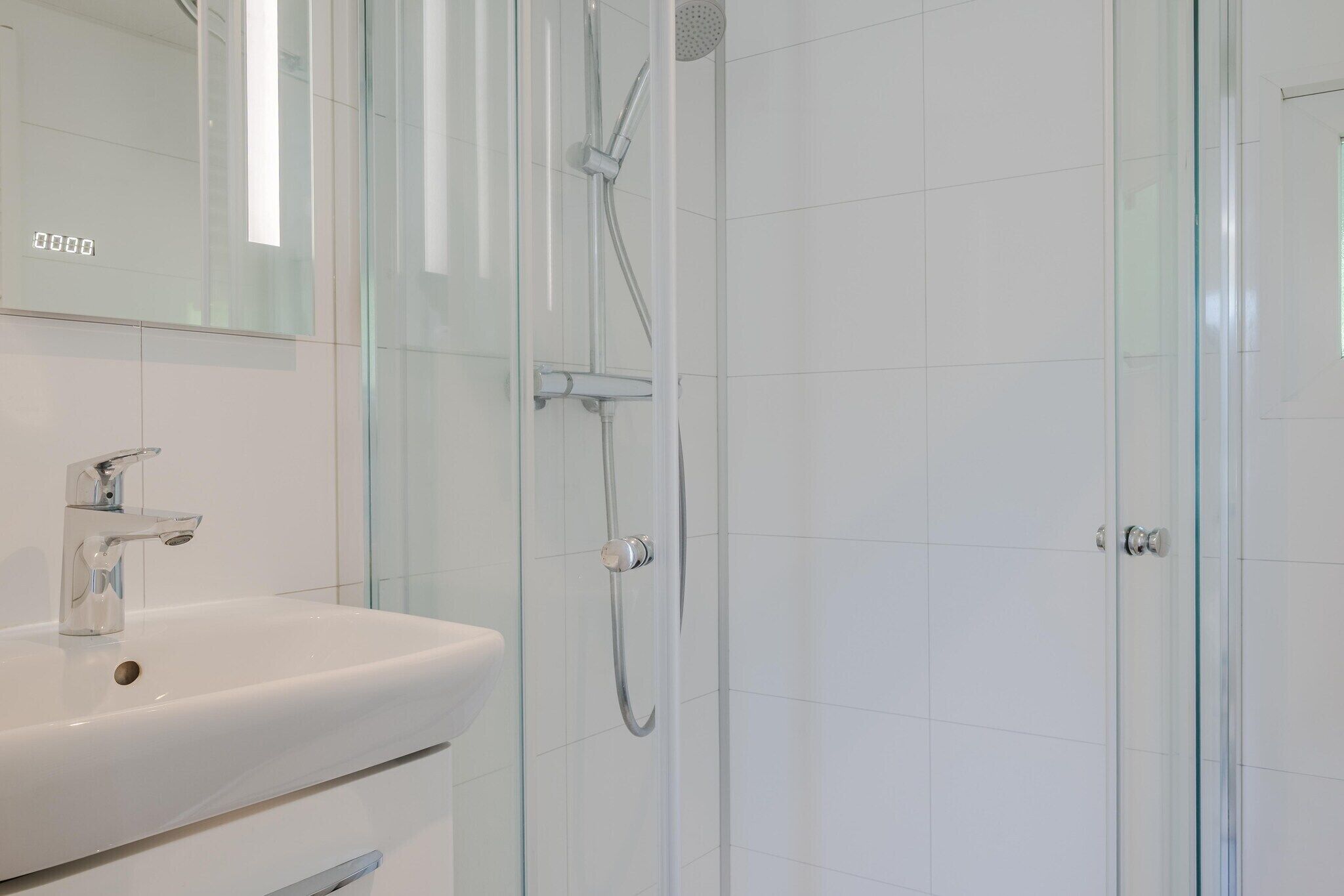 Ein modernes Badezimmer mit Waschbecken in Weiß, einer Duschkabine aus Glas und einer Tür mit Griff.