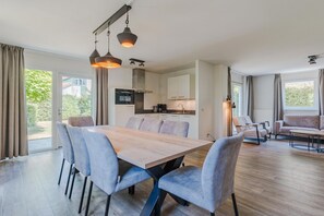 A modern dining area with a wooden table and grey upholstered chairs, a kitchen in the background, and large windows with curtains. - Nature-Set Villa with Microwave Oven (Arcen)