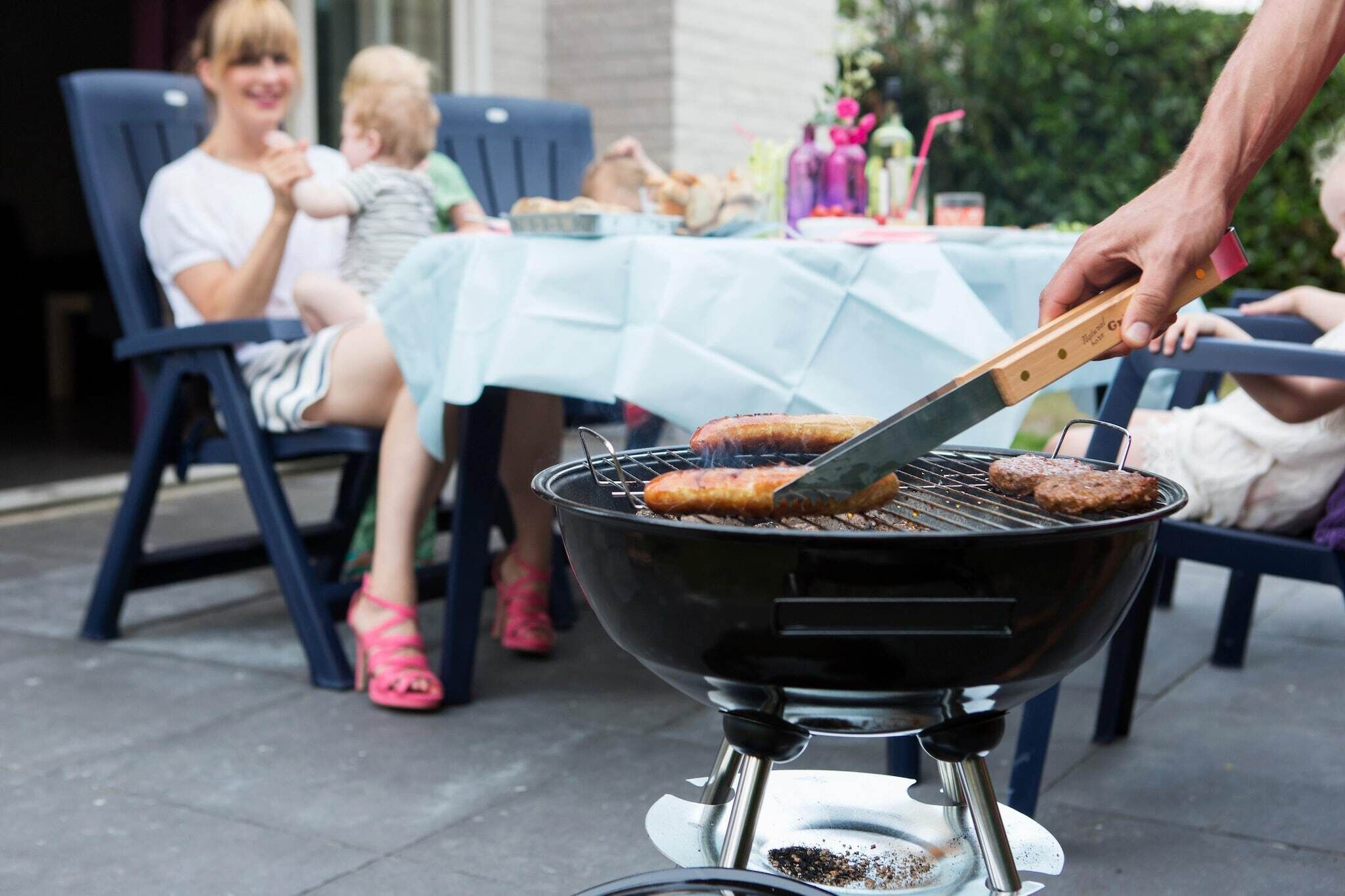 Una parrilla con dos hamburguesas asándose, una mesa con mantel azul claro y personas sentadas alrededor.