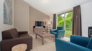 A modern living room with a sofa, armchair, and coffee table. A flat-screen TV is mounted on the wall, and there is a window with curtains. - Modern Bungalow with Washer (Arcen)