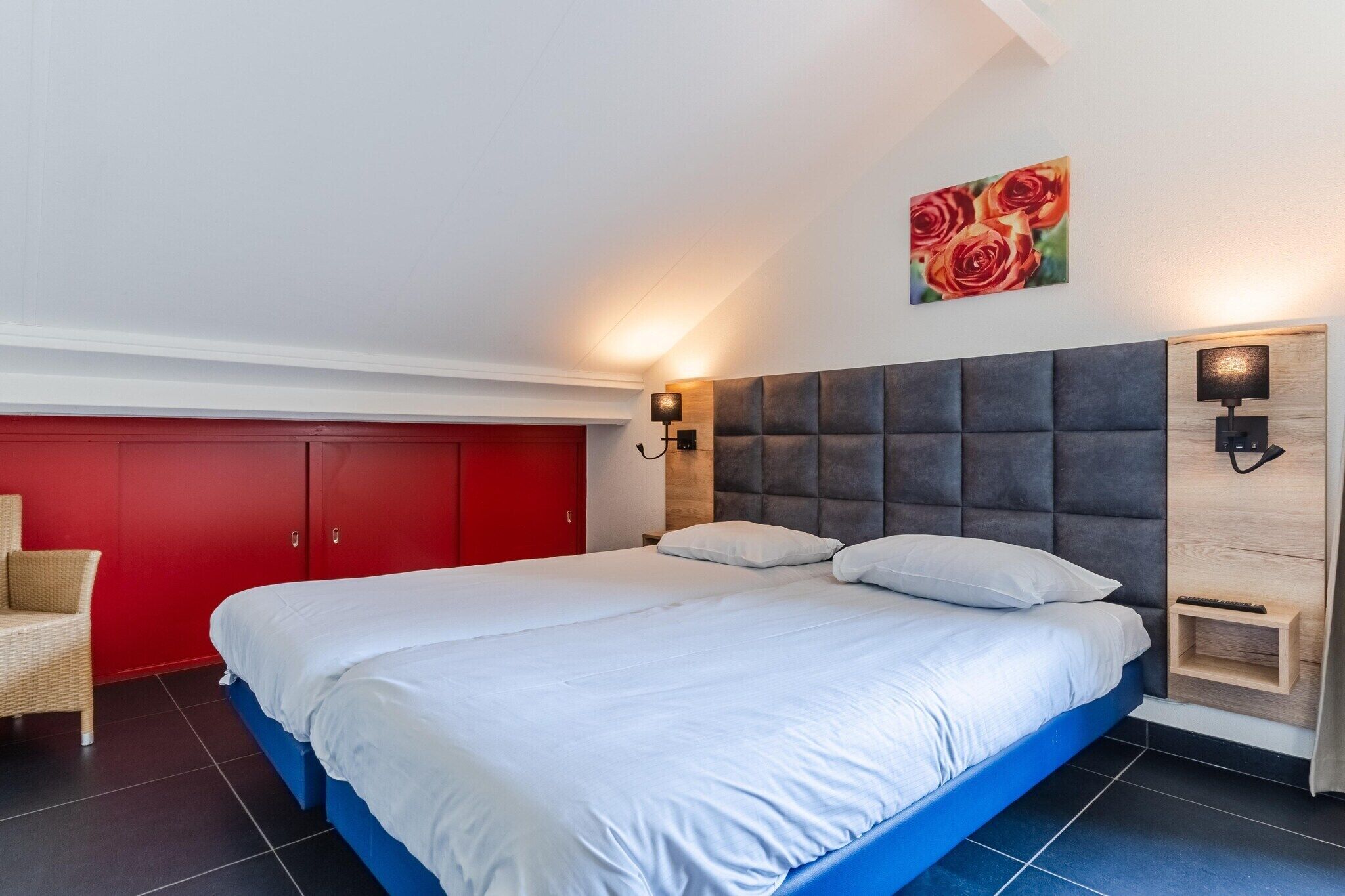 Une chambre à coucher avec un lit, une armoire rouge, une lampe fixée au mur et un tableau représentant des roses.