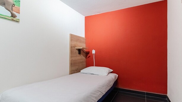 Una habitación pequeña con cama individual, pared de acento roja, cabecero de madera y almohada blanca.