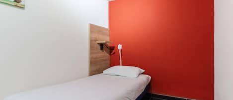 A small, single-bed room with a red accent wall, a wooden headboard, and a white pillow.
