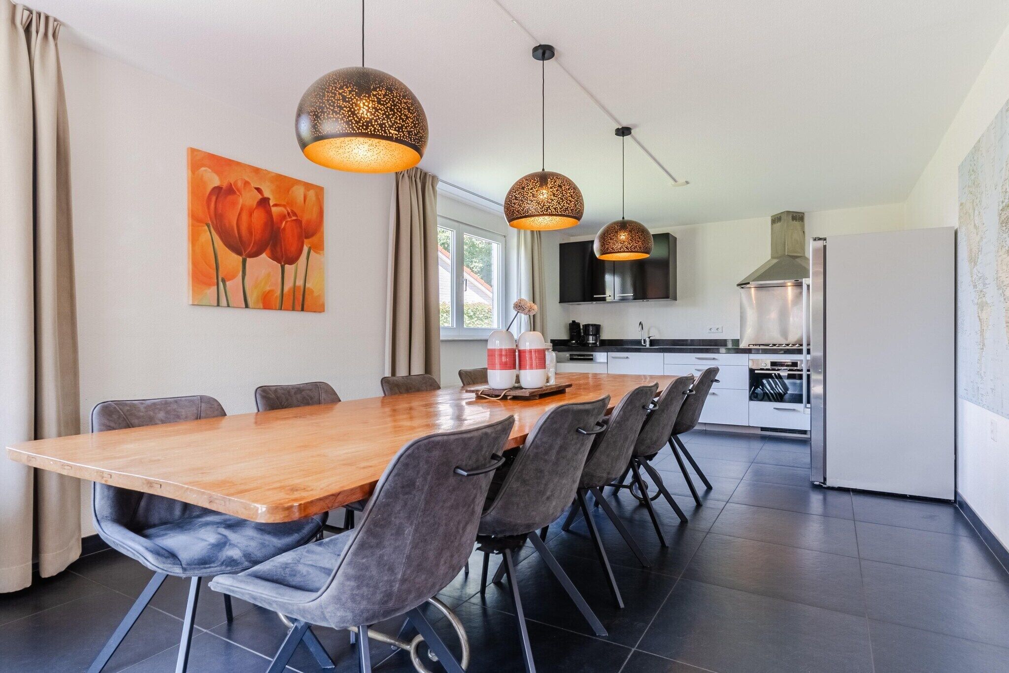 A modern kitchen with a large dining table, grey upholstered chairs, pendant lighting, and a painting of tulips on the wall.