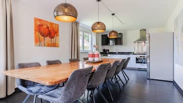A modern kitchen with a large dining table, grey upholstered chairs, pendant lighting, and a painting of tulips on the wall.