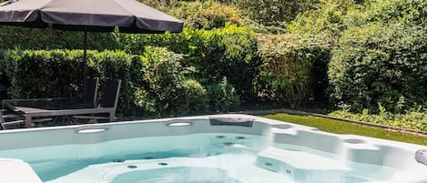 A hot tub with a patio chair and umbrella, surrounded by lush greenery.