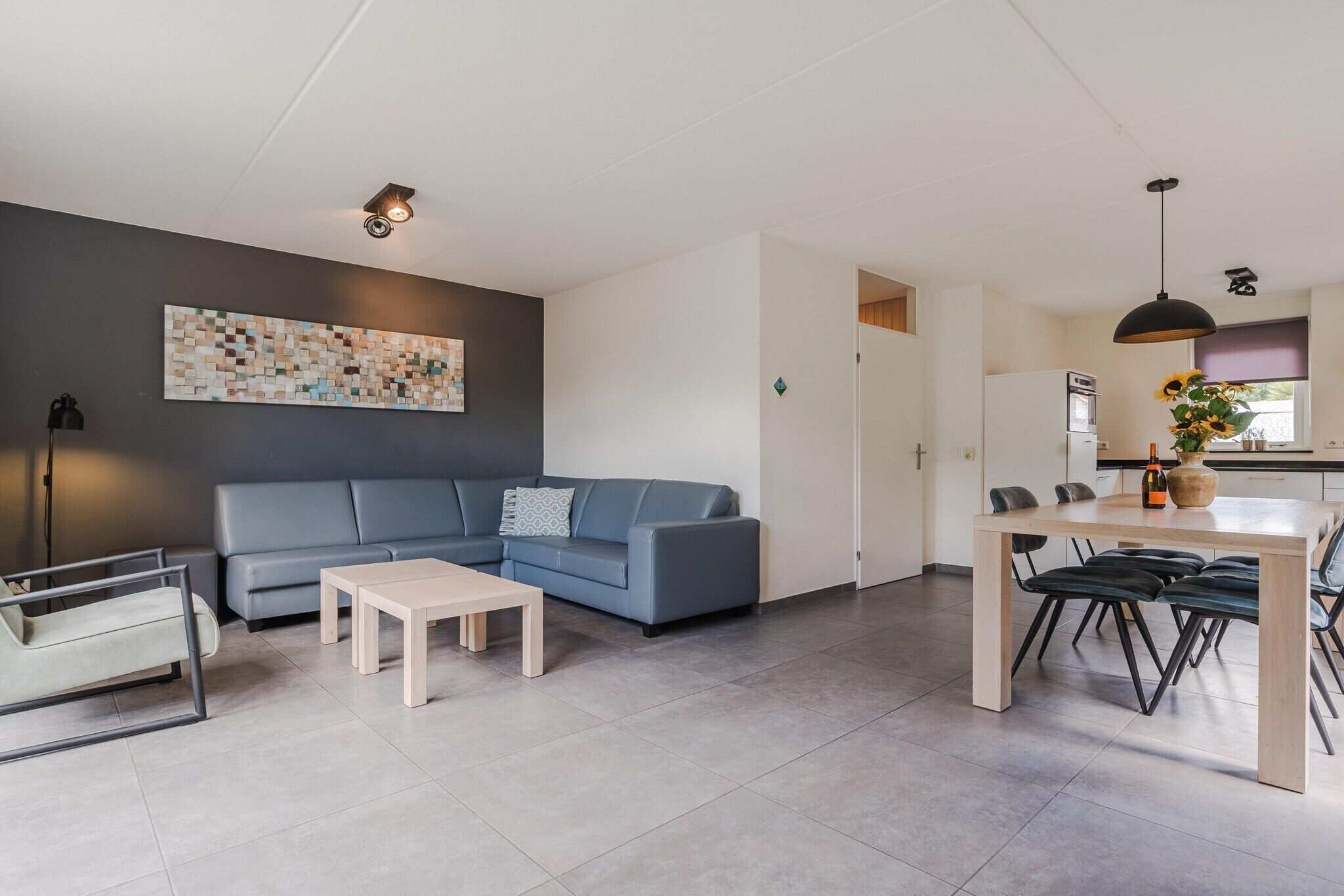 A modern living room with a blue sofa, a coffee table, a dining area with a table and chairs, and a kitchen visible through an open door.