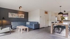 Moderni olohuone, jossa on sininen sohva, sohvapöytä, ruokailutila pöytineen ja tuoleineen sekä avoimen oven kautta näkyvä keittiö.