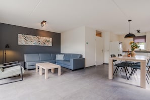 A modern living room with a blue sofa, a coffee table, a dining area with a table and chairs, and a kitchen visible through an open door.