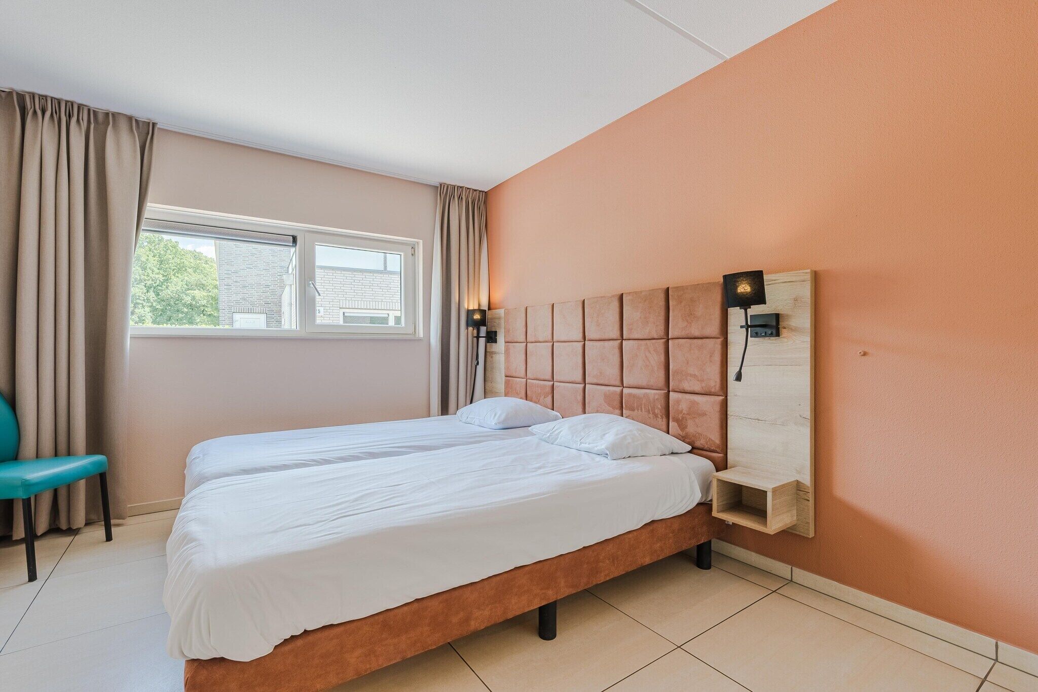Ein Doppelbett mit Holz-Kopfteil, ein Nachttisch und ein Stuhl in einem Zimmer mit beige gestrichenen Wänden und einem fenster mit Vorhängen.