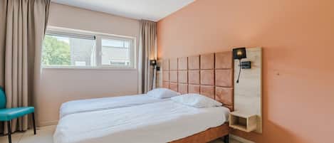 A double bed with a wooden headboard, a bedside table, and a chair in a room with beige walls and a window with curtains.