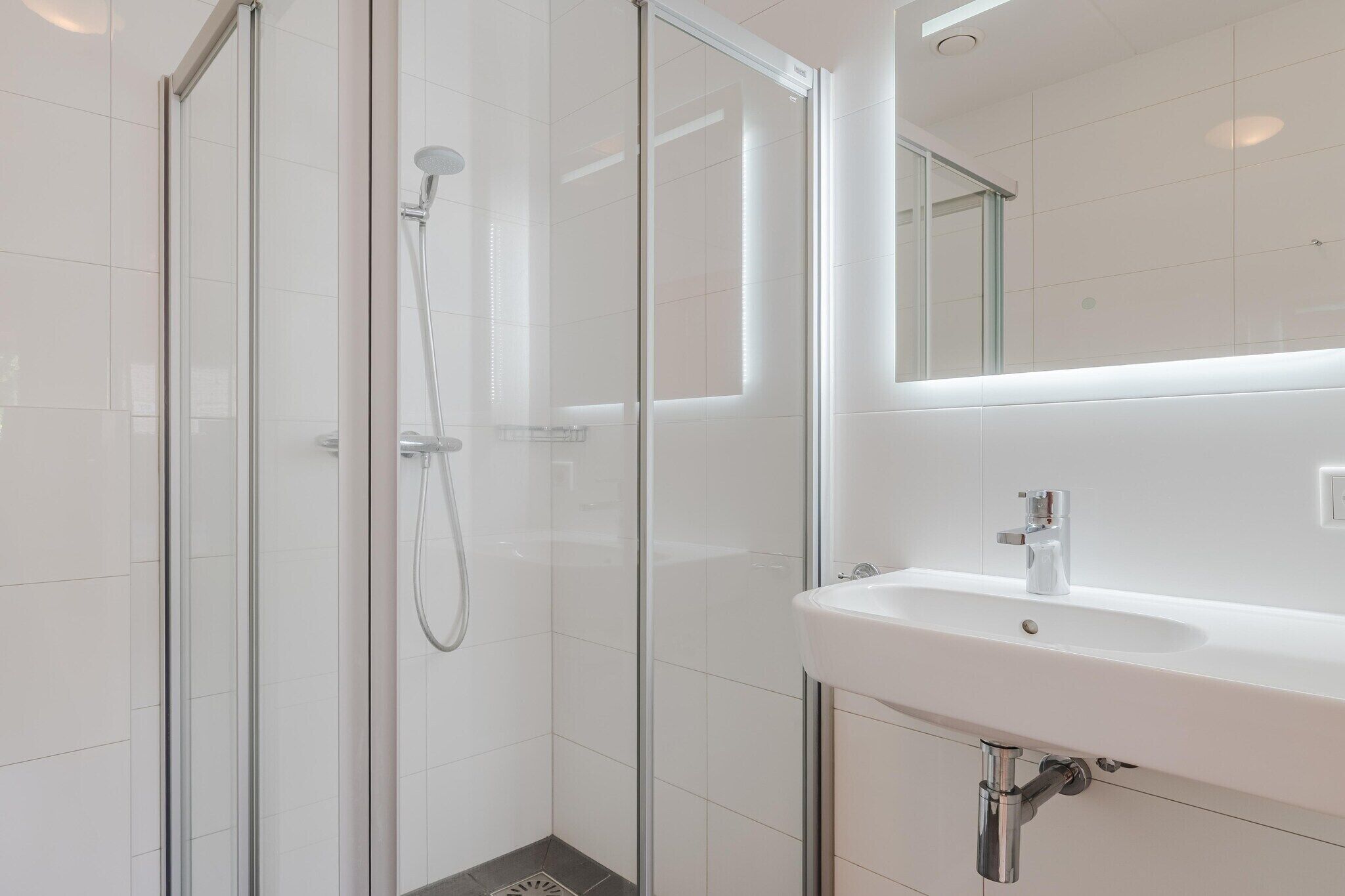 Een moderne badkamer met een douche in glazen cabine, een witte wastafel en een spiegel erboven.