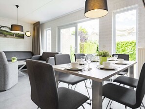 A modern dining area with a rectangular table set for six, surrounded by six chairs. There are two pendant lights above the table, and the room has large windows providing a view of greenery outside. - Cottage in Gelderland with Fenced Garden (Plasmolen)