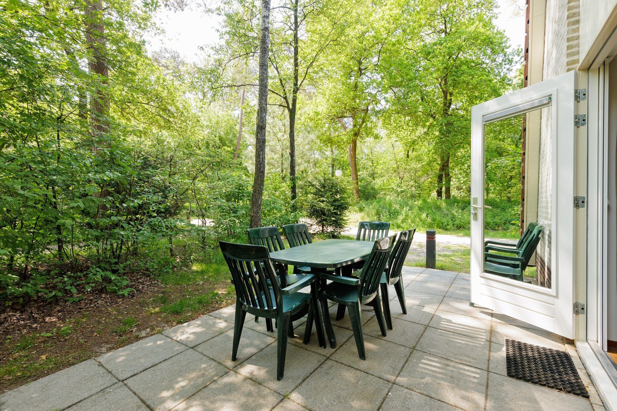 Une terrasse extérieure avec une table et des chaises vertes, entourée d’arbres et d’un bâtiment blanc.