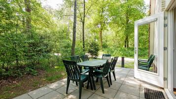 Une terrasse extérieure avec une table et des chaises vertes, entourée d’arbres et d’un bâtiment blanc.