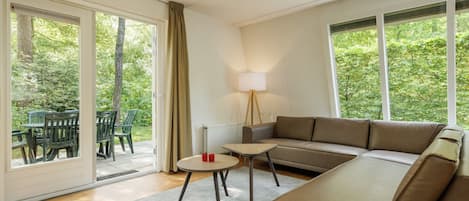 A modern living room with a brown sofa, a small round table, and a lamp. There is a sliding glass door leading to an outdoor area with chairs and a table.