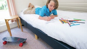 Un enfant qui dessine sur un lit avec des crayons de couleur, un skateboard et un petit tabouret en bois.